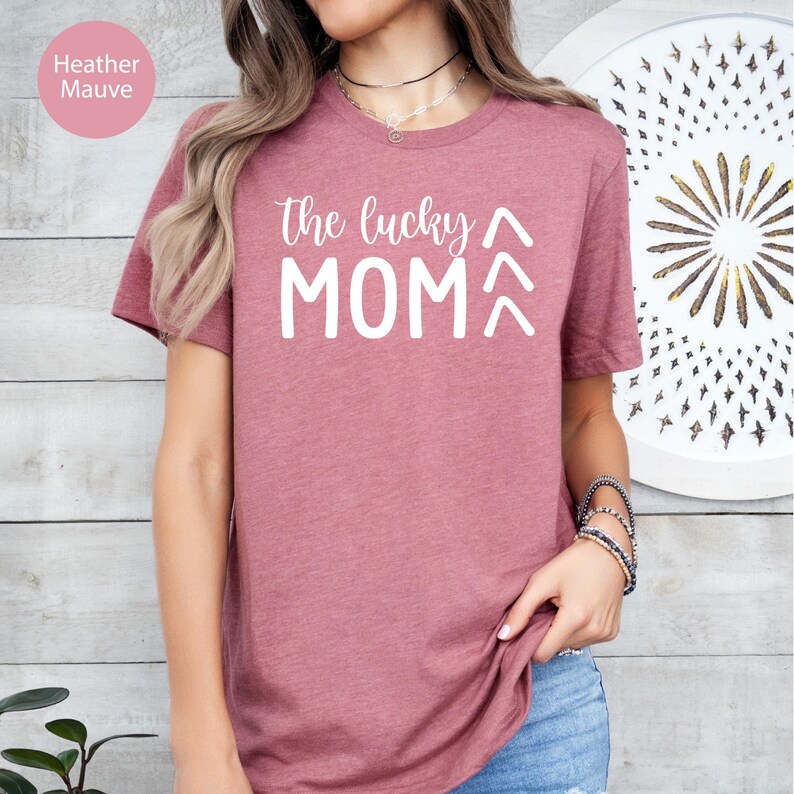 May include: Heather mauve t-shirt with the text "the lucky MOM" in white. The shirt has short sleeves and a crew neck. The text is in a handwritten style font.