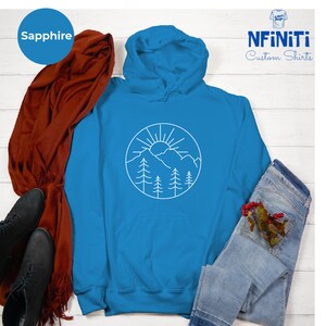 K&ouml;nnte beinhalten: Ein saphiblau-farbener Hoodie mit einem wei&szlig;en Umriss einer Sonne, Bergen und B&auml;umen in einem Kreis. Der Hoodie hat eine Kapuze mit Kordelzug und eine K&auml;ngurutasche.
