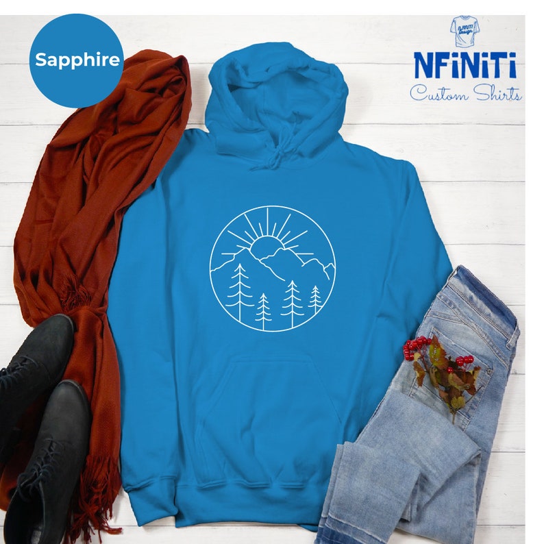 K&ouml;nnte beinhalten: Ein saphiblau-farbener Hoodie mit einem wei&szlig;en Umriss einer Sonne, Bergen und B&auml;umen in einem Kreis. Der Hoodie hat eine Kapuze mit Kordelzug und eine K&auml;ngurutasche.