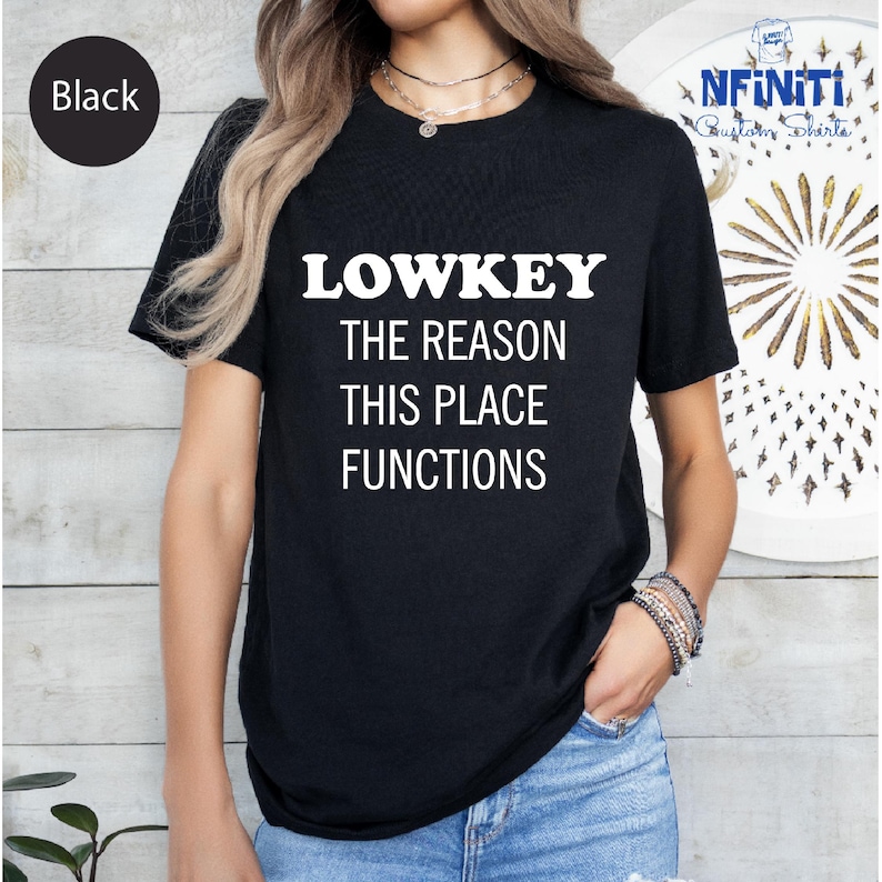 A person wearing a black t-shirt with white text that says "LOWKEY THE REASON THIS PLACE FUNCTIONS".