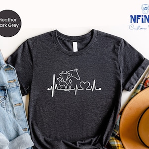 May include: Heather dark grey t-shirt with a white outline of a horse, cow, and calf with a heartbeat and heart graphic. The text "NFINITI Custom Shirts" is in the top right corner of the image.