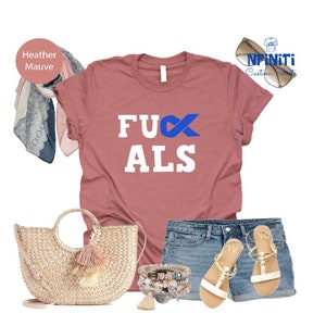 May include: A heather mauve t-shirt with the text "FUX ALS" in white letters with a blue ribbon. The shirt is being modeled with a straw bag, denim shorts, and white sandals.