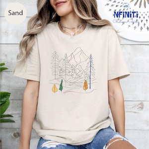 Peut inclure: Un t-shirt beige avec un dessin au trait d'une chaîne de montagnes et d'arbres. Le dessin est dans des tons de vert, jaune, orange et bleu.