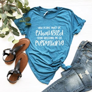 May include: A teal blue t-shirt with the text "You people must be exhausted from watching me do everything" in white.