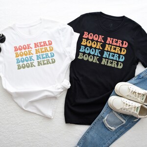 May include: Two t-shirts with the text "BOOK NERD" printed in a rainbow color scheme. The shirts are laid out on a white surface with a pair of white sneakers and blue jeans.