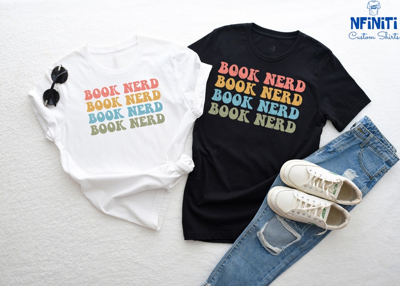 May include: Two t-shirts with the text "BOOK NERD" printed in a rainbow color scheme. The shirts are laid out on a white surface with a pair of white sneakers and blue jeans.