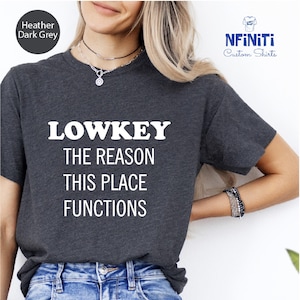 A woman wearing a gray t-shirt with the text "LOWKEY THE REASON THIS PLACE FUNCTIONS" printed on it.