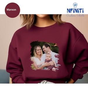 May include: A maroon sweatshirt with a photo of a family of three printed on the front. The photo is in a white frame with a brushstroke effect. The text "NFINITI Custom Shirts" is printed in blue on the top right corner of the sweatshirt.