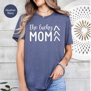 May include: Heather navy blue t-shirt with the text "the lucky MOM" in white. The shirt has short sleeves and a crew neck. The text includes two upward-pointing arrow symbols above the word "MOM".