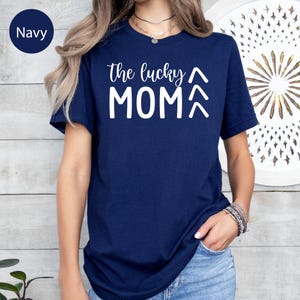 May include: Navy blue t-shirt with the text "the lucky MOM" in white. The shirt has short sleeves and a crew neck. The text includes an upward arrow above the word "MOM".