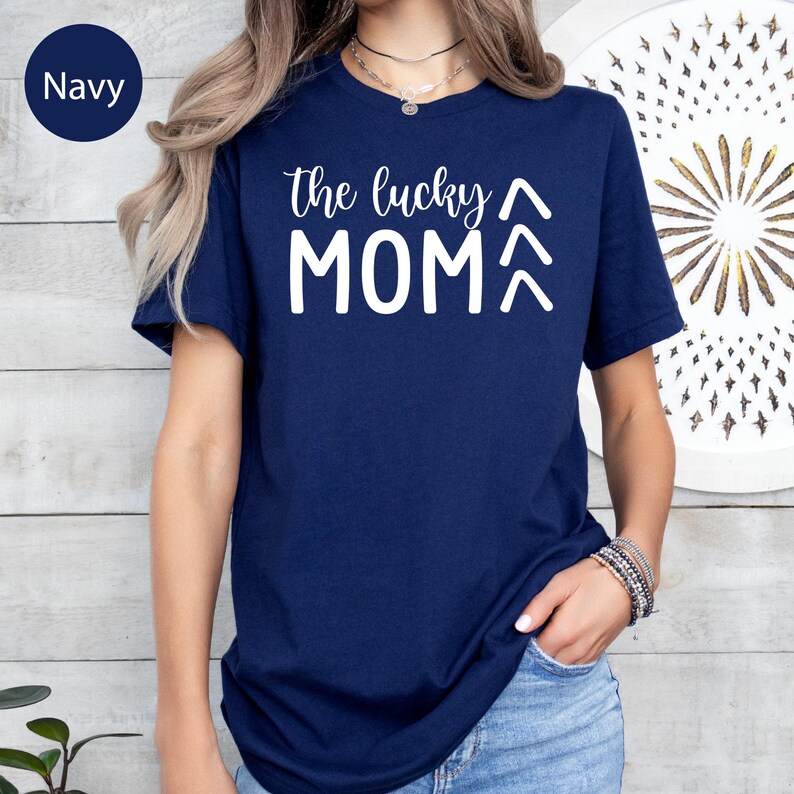 May include: Navy blue t-shirt with the text "the lucky MOM" in white. The shirt has short sleeves and a crew neck. The text includes an upward arrow above the word "MOM".