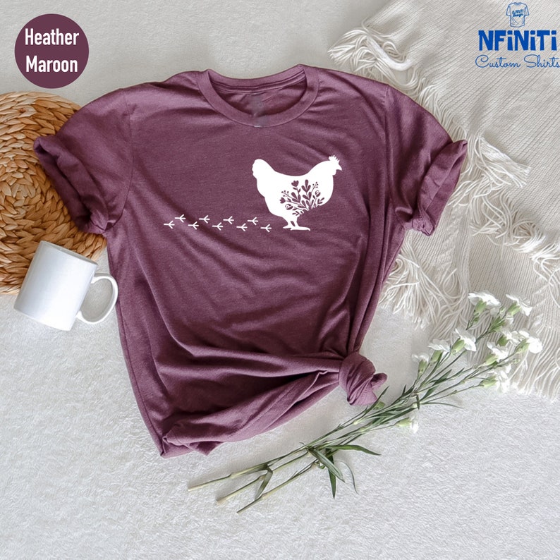 May include: A heather maroon t-shirt with a white silhouette of a chicken with floral details and a trail of footprints behind it. The shirt is made of a soft, comfortable material and is perfect for everyday wear.