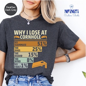 Pode incluir: Uma camiseta cinza escuro heather com um gráfico de estilo vintage que diz "Why I Lose at Cornhole" e lista as razões para perder no jogo com porcentagens. O gráfico inclui uma ilustração estilizada de um tabuleiro de cornhole.