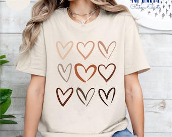 Pode incluir: Uma camiseta bege com um gráfico de nove corações de diferentes cores. Os corações estão dispostos em três filas de três. A camiseta é de corte relaxado e tem mangas curtas.
