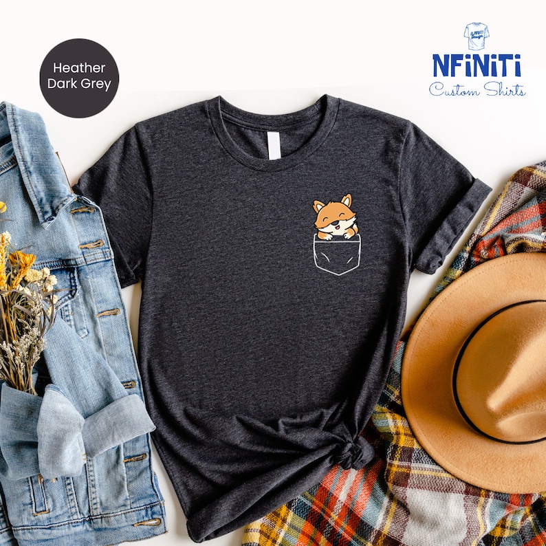 May include: A heather dark grey t-shirt with a small pocket on the front. A cartoon fox is peeking out of the pocket. The shirt is laid flat on a white background with a denim jacket, a plaid blanket, and a brown hat.
