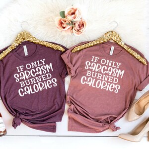 May include: Two heather maroon t-shirts with the text "If only sarcasm burned calories" on a white background. The shirts are hanging on gold hangers.