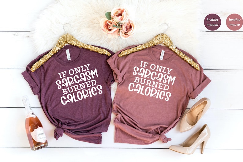 May include: Two heather maroon t-shirts with the text "If only sarcasm burned calories" on a white background. The shirts are hanging on gold hangers.