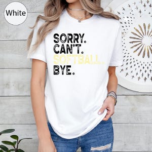 May include: White t-shirt featuring the text "SORRY. CAN'T. SOFTBALL. BYE." in black and yellow. The shirt has a crew neck and is worn by a person. The t-shirt is a casual wear item.