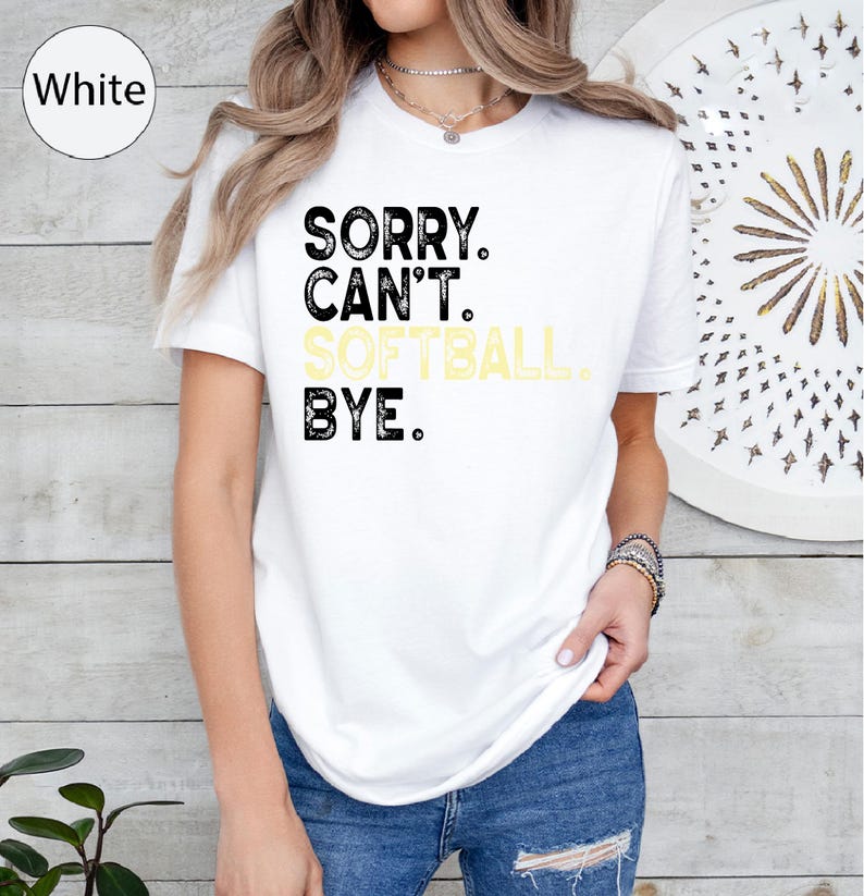 May include: White t-shirt featuring the text "SORRY. CAN'T. SOFTBALL. BYE." in black and yellow. The shirt has a crew neck and is worn by a person. The t-shirt is a casual wear item.