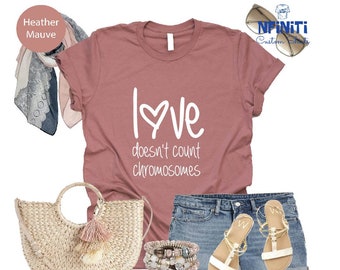 May include: A heather mauve t-shirt with the text "Love doesn't count chromosomes" in white. The shirt is styled with denim shorts, a straw bag, and sandals.