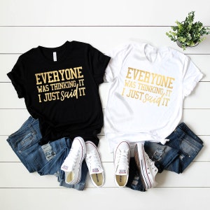 Pode incluir: Duas camisetas com o texto "Everyone was thinking it, I just said it" em letras douradas em uma camiseta preta e branca. As camisetas est&atilde;o dispostas em uma superf&iacute;cie de madeira branca com jeans azul e t&ecirc;nis brancos.