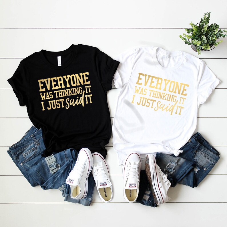 Pode incluir: Duas camisetas com o texto "Everyone was thinking it, I just said it" em letras douradas em uma camiseta preta e branca. As camisetas est&atilde;o dispostas em uma superf&iacute;cie de madeira branca com jeans azul e t&ecirc;nis brancos.