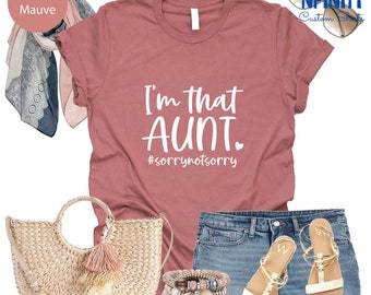 Puede incluir: Una camiseta color malva jaspeado con el texto blanco "I'm that AUNT. #sorrynotsorry". El conjunto incluye shorts vaqueros, sandalias, un bolso de paja, pulseras y una bufanda.
