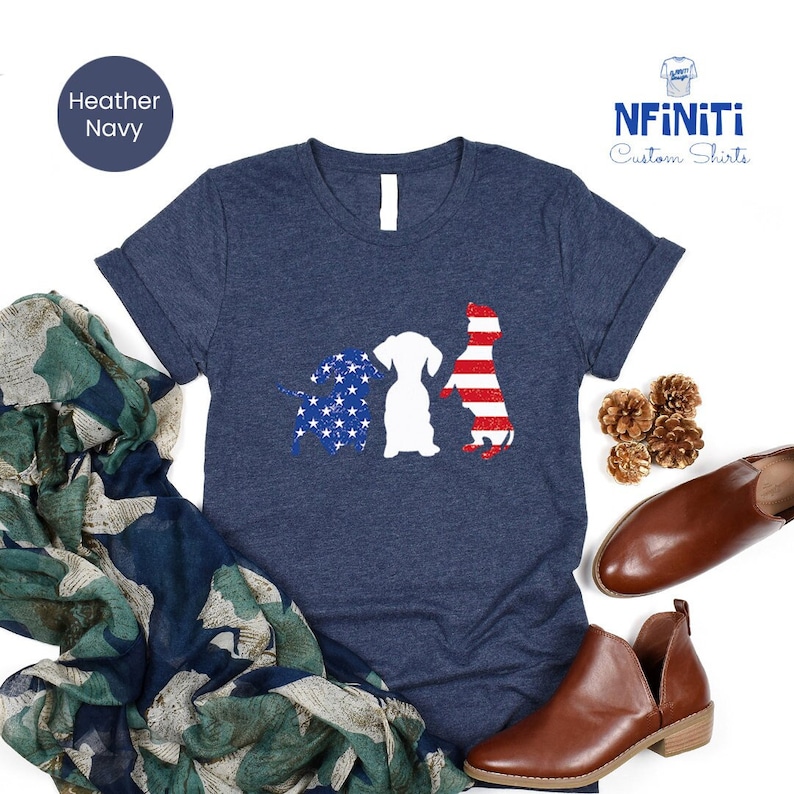 May include: A heather navy blue t-shirt with a design of three dogs in silhouette. The dogs are outlined in red, white, and blue, resembling the American flag. The text "NFINITI Custom Shirts" is in the top right corner of the image.