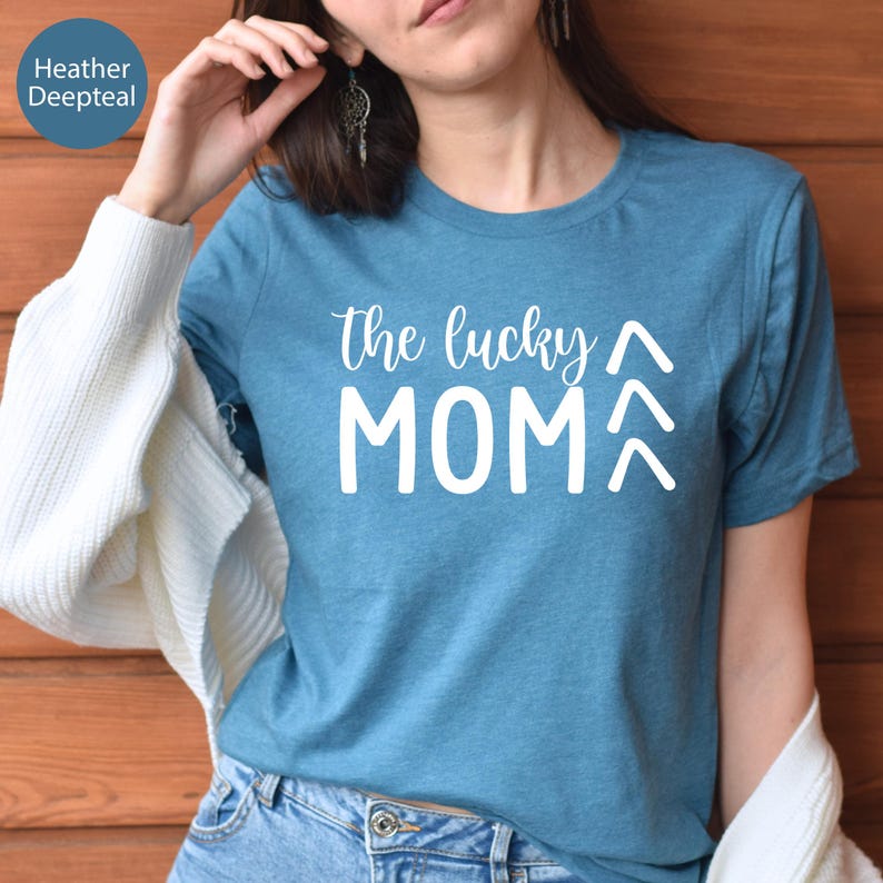 May include: A teal t-shirt with the text "the lucky MOM" in white script and block letters. Two upward-pointing arrows are below the word MOM. The shirt is made of a soft material.