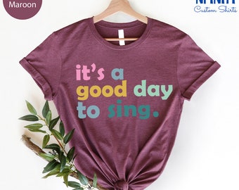 May include: Heather maroon t-shirt with the text "it's a good day to sing." in colorful letters.