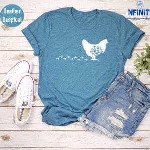 May include: A heather deepteal t-shirt with a white silhouette of a chicken with floral details and a trail of footprints. The shirt is laid flat on a white wooden surface with a pair of white sneakers and a pair of denim shorts.