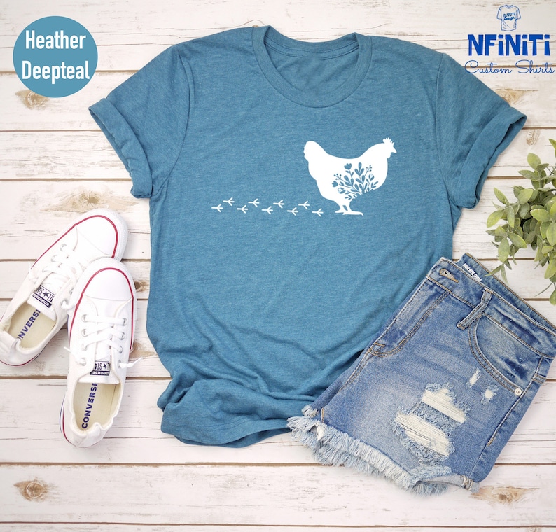 May include: A heather deepteal t-shirt with a white silhouette of a chicken with floral details and a trail of footprints. The shirt is laid flat on a white wooden surface with a pair of white sneakers and a pair of denim shorts.