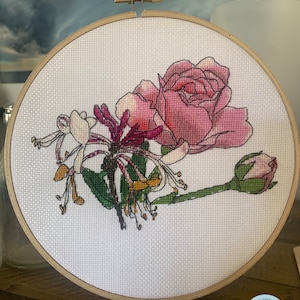 May include: A white embroidery hoop with a pink rose and white honeysuckle flowers embroidered on it. The hoop is made of wood and has a brass screw on the top. The embroidery is signed "Cotton Tree Studios, Created for You".
