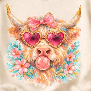 May include: A cream-coloured sweatshirt with a playful design of a Highland cow wearing heart-shaped sunglasses and a pink bow. The cow is blowing a bubblegum bubble, surrounded by flowers. The design is printed on a soft, comfortable fabric.