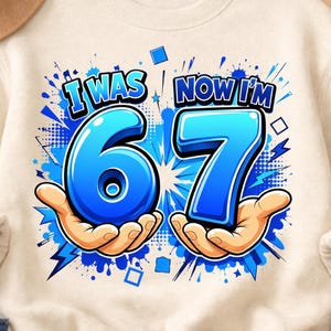 May include: A cream-colored crewneck sweatshirt featuring a graphic print with the text I WAS 67 NOW I'M 7. The design includes large blue numbers 6 and 7, each held by a cartoon hand, surrounded by blue comic-style splashes, dots, and squares.
