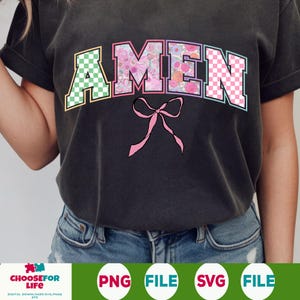 Op de afbeelding: Een zwart T-shirt met het woord "AMEN" in kleurrijke letters met bloemen- en ruitpatronen. Onder het woord zit een roze strik.
