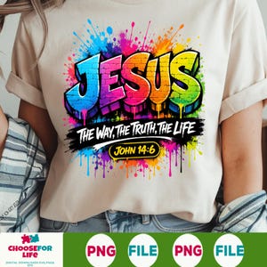 May include: A beige t-shirt featuring a colorful graphic with the word JESUS in a brick-patterned font, surrounded by paint splatters. Below, the text reads THE WAY, THE TRUTH, THE LIFE and JOHN 14:6 in a black brushstroke design with yellow accents.