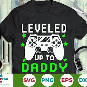Può includere: Maglietta nera con una grafica di un controller di videogiochi bianco e il testo "LEVELED UP TO DADDY" in lettere pixelate verdi.