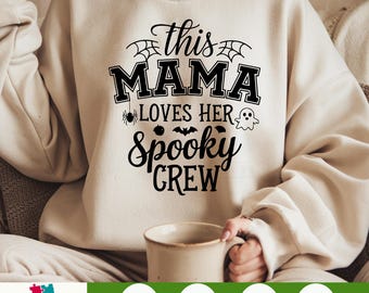 Könnte beinhalten: Cremefarbenes Sweatshirt mit dem schwarzen Text "this MAMA loves her Spooky CREW". Das Design umfasst Spinnweben, eine Spinne, eine Fledermaus und einen Geist. Eine Tasse wird in der Hand der Person gehalten.