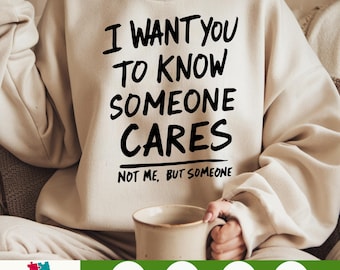 May include: Cream-colored sweatshirt with the text "I WANT YOU TO KNOW SOMEONE CARES" in black, with "NOT ME, BUT SOMEONE" underneath. The person is holding a beige mug. The bottom left corner has a logo that says "CHOOSEFOR LIFE".
