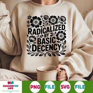 Op de afbeelding: Crèmekleurige sweatshirt met de zwarte tekst "RADICALIZED BY BASIC DECENCY", omringd door bloemen- en bladmotieven. Het ontwerp is gecentreerd op de voorkant van de top met lange mouwen. Een koffiemok is ook zichtbaar.