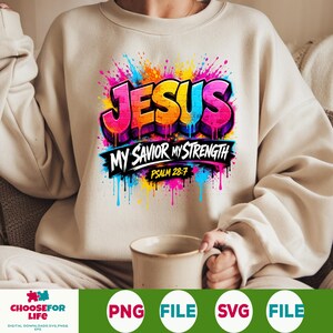 May include: A beige crewneck sweatshirt featuring a vibrant, multi-colored graphic print. The design displays the word JESUS in a graffiti-style font with paint drips, followed by the text MY SAVIOR MY STRENGTH and PSALM 28:7 in black.