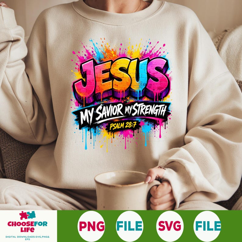 May include: A beige crewneck sweatshirt featuring a vibrant, multi-colored graphic print. The design displays the word JESUS in a graffiti-style font with paint drips, followed by the text MY SAVIOR MY STRENGTH and PSALM 28:7 in black.