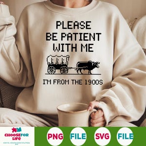 Op de afbeelding: Een beige sweatshirt met de tekst "PLEASE BE PATIENT WITH ME I'M FROM THE 1900S" in zwart. Onder de tekst staat een zwart-wit gepixelde afbeelding van een gedekte wagen die door een os wordt getrokken.