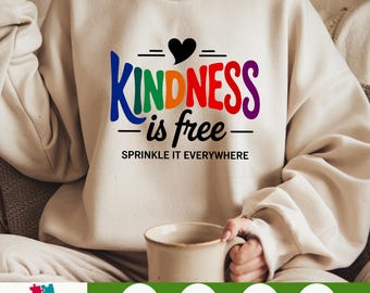 Può includere: Felpa color crema con le parole "KINDNESS is free" in colori arcobaleno e un cuore nero. La frase "SPRINKLE IT EVERYWHERE" è sotto il testo principale. Una tazza è tenuta in mano.