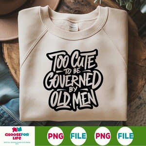 Peut inclure: Un sweat-shirt crème avec le texte "Too Cute to be Governed by Old Men" en noir et blanc. Le sweat-shirt est présenté sur une surface en bois.