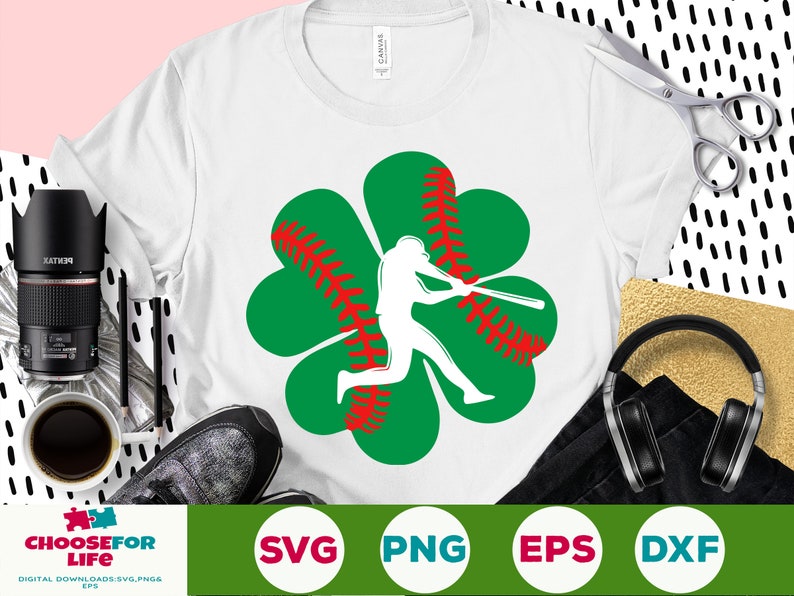 May include: A white t-shirt with a green four-leaf clover design. The clover has a red baseball stitch pattern and a silhouette of a baseball player swinging a bat.