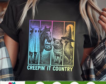 May include: Black t-shirt with a Halloween-themed graphic. The design features a cow in a ghost costume, a horse with a jack-o'-lantern, a pig, and a chicken wearing a witch hat. The text "CREEPIN' IT COUNTRY" is below the image.