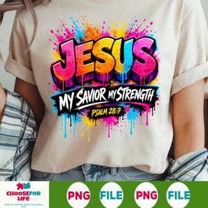 May include: A beige t-shirt featuring a colorful graphic with the word JESUS in a brick-patterned font, surrounded by vibrant paint splatters. Below, the text reads MY SAVIOR MY STRENGTH and PSALM 28:7 in a bold, black, dripping graffiti style.