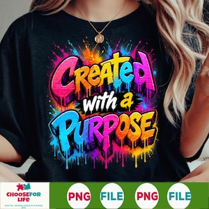May include: A black t-shirt featuring a vibrant, multi-colored graffiti-style graphic with the text "CREATED with a PURPOSE". The design incorporates bright pink, orange, yellow, and blue paint splatters and drips over a brick-like texture.